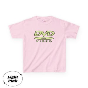 May include: A light pink short-sleeved t-shirt featuring a graphic on the chest with the text DVD VIDEO in a stylized lime green font. The design includes a circular disc shape beneath the letters. The shirt has a crew neckline and short sleeves.