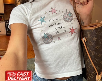 May include: A white short-sleeved t-shirt featuring a graphic print of two disco balls and several stars in pink and light blue. The text "IT'S ALL WAITING THERE FOR YOU" is printed in black in the center of the design, surrounded by small dots.