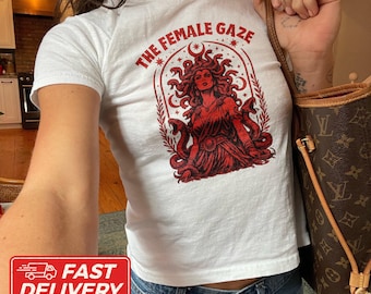 May include: A white short-sleeved t-shirt featuring a red graphic print of a figure with snakes for hair, surrounded by an arched frame and stars. The text THE FEMALE GAZE is printed in a red arc above the illustration on the front of the shirt.