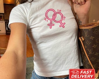 May include: A white short-sleeved t-shirt featuring a graphic of two interlocking pink female gender symbols. The design includes small pink sparkles and hearts surrounding the symbols. The shirt is made of cotton and is shown in a fitted style.
