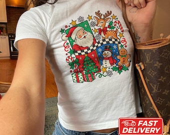 May include: A white short-sleeved t-shirt featuring a colorful Christmas graphic print. The design includes Santa Claus, a reindeer, a snowman, a gingerbread man, a decorated tree, gift boxes, candy canes, and stars on a checkered background.