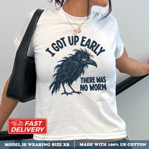 Może przedstawiać: Biały t-shirt z krótkim rękawem z ciemnoniebieską grafiką przedstawiającą zrzędliwego kruka. Tekst nad krukiem brzmi I GOT UP EARLY, a tekst pod krukiem brzmi THERE WAS NO WORM. Koszulka jest noszona z czarną torbą na ramię.