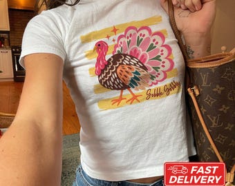 May include: A white short-sleeved t-shirt featuring a graphic of a turkey with pink and brown feathers, yellow horizontal brush strokes, and the text Gobble Gobble in cursive script. The shirt is made of cotton and has a crew neckline.