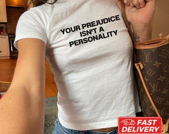 May include: A white short-sleeved t-shirt featuring the black text "YOUR PREJUDICE ISN'T A PERSONALITY" printed on the chest. The shirt is paired with blue denim jeans and a brown patterned shoulder bag with gold-tone hardware.