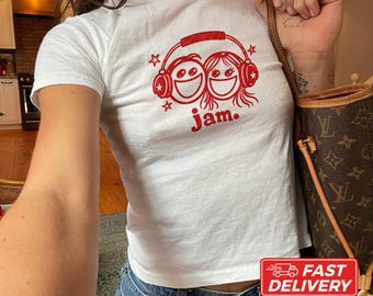 May include: A white short-sleeved t-shirt featuring a red graphic of two smiling faces wearing headphones with the text jam. below the design. The shirt is made of cotton and is shown in a size XS. The design includes small star accents.