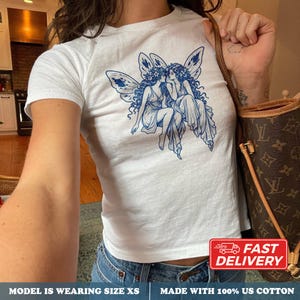 Puede incluir: Una camiseta blanca de manga corta con un estampado gráfico azul de dos figuras aladas con cabello largo y prendas fluidas. La camiseta está hecha de 100% algodón y se muestra en talla XS. Se ve una correa de bolso marrón con un patrón.