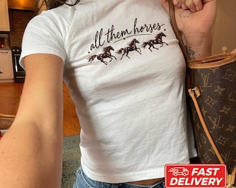 May include: A white short-sleeved t-shirt featuring the black cursive text all them horses above a graphic of three galloping horses. The shirt is made of cotton and is shown in a size XS. The design includes small stars between the words.