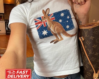 May include: A white short-sleeved t-shirt featuring a graphic of a brown kangaroo standing in front of a blue, red, and white Australian flag. The design appears to be rendered in a textured, crayon-like style on the front of the cotton garment.