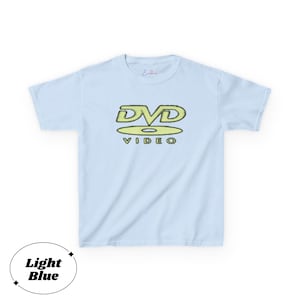 May include: A light blue short-sleeve t-shirt featuring a graphic on the chest. The graphic displays the text DVD in a stylized yellow font above a yellow disc icon and the word VIDEO in a smaller yellow font. The shirt has a crew neckline.