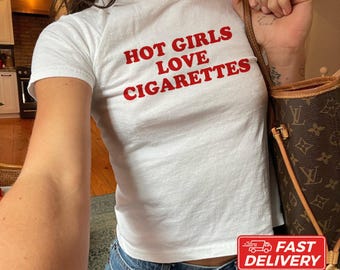 May include: A white short-sleeved t-shirt featuring the red text HOT GIRLS LOVE CIGARETTES printed on the chest. The shirt is paired with a brown monogrammed shoulder bag and blue denim jeans. The garment has a crew neckline.