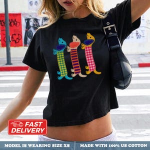 Può includere: Una t-shirt nera a maniche corte con una stampa grafica di tre cani bassotti stilizzati. Ogni cane presenta un motivo diverso sul corpo: uno a quadri gialli e blu, uno a righe arancioni e viola e uno a quadretti vichy rosa e bianchi.