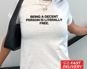 May include: A white short-sleeved t-shirt featuring the black text "BEING A DECENT PERSON IS LITERALLY FREE." The shirt is paired with a black shoulder bag and a gold-toned necklace with a small circular pendant.