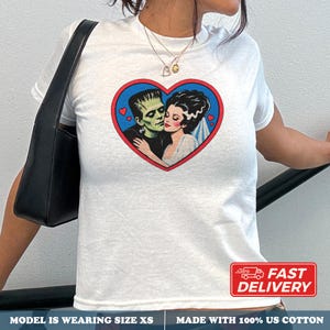 Pode incluir: Uma t-shirt branca de manga curta com uma estampa gráfica do monstro de Frankenstein e a sua noiva dentro de um coração azul com rebordo vermelho. O design inclui pequenos corações vermelhos a rodear o casal num fundo branco.