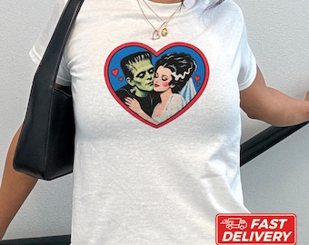 May include: A white short-sleeved t-shirt featuring a graphic print of Frankenstein's monster and his bride inside a red-bordered blue heart. The design includes small red hearts surrounding the couple, set against a plain white background.