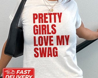 May include: A white short-sleeved t-shirt featuring the red text PRETTY GIRLS LOVE MY SWAG printed on the front. The shirt has a crew neckline and is paired with a black shoulder bag. The design is centered on the chest in a bold, sans-serif font.