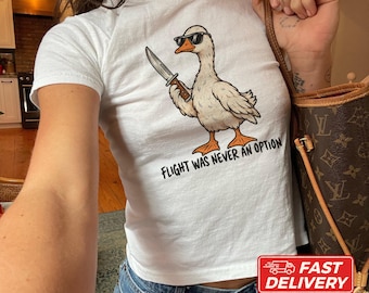 May include: A white short-sleeved t-shirt featuring a graphic of a white duck wearing black sunglasses and holding a knife in its wing. Below the duck, the text reads, "FLIGHT WAS NEVER AN OPTION" in black capital letters.