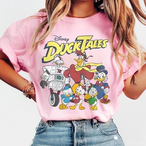 Puede incluir: Una camiseta rosa claro con un estampado gráfico del logotipo de Disney DuckTales en texto amarillo y negro, acompañado de ilustraciones de los personajes Gizmoduck, Launchpad McQuack, Scrooge McDuck y los tres patitos Huey, Dewey y Louie.