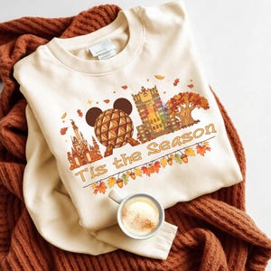 May include: A cream-coloured crewneck sweatshirt featuring a graphic print of a geodesic dome, a castle, and a tall building, with the text 'Tis the Season' written in a cursive font, surrounded by autumn leaves, acorns, and small stars.