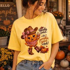 Puede incluir: Una camiseta amarilla con un gráfico de un personaje de calabaza sonriente que lleva un sombrero de vaquero, botas y un pañuelo, sosteniendo una taza de café. El texto dice: Howdy, partner it's pumpkin spice o'clock! con corazones y hojas.
