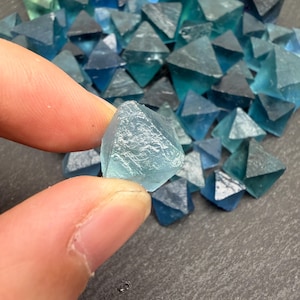 May include: A collection of small, translucent blue fluorite crystals, each shaped as a natural octahedron. The stones exhibit varying shades of teal and deep blue, with a glassy, crystalline texture and sharp geometric edges throughout the pile.