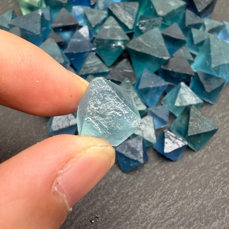 May include: A collection of small, translucent blue fluorite crystals, each shaped as a natural octahedron. The stones exhibit varying shades of teal and deep blue, with a glassy, crystalline texture and sharp geometric edges throughout the pile.