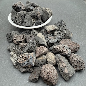 Peut inclure: Une collection de roches volcaniques poreuses gris foncé et noires. Certaines pierres sont contenues dans un bol circulaire blanc, tandis que d'autres sont éparpillées en tas. Chaque pierre présente une texture rugueuse avec des trous visibles.