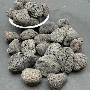 以下が含まれることがあります： 暗い灰色の多孔質な火山溶岩石のコレクション。いくつかの石は小さな白いボウルの中に入っており、他の石はボウルの底の周りに散らばっています。それぞれの石は、目に見える穴がある粗い、くぼんだ質感の特徴を持っています。