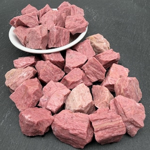 May include: A collection of raw, dusty pink rhodonite mineral chunks. Some stones are piled inside a round white ceramic bowl, while others are scattered around the base of the bowl, displaying natural jagged edges and crystalline textures.