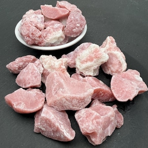 May include: A collection of raw pink rhodonite stones with white mineral inclusions. Some stones are piled inside a small white bowl, while others are scattered around the base of the bowl, displaying irregular shapes and a matte, textured surface.