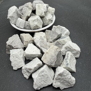 May include: A collection of rough, light grey and white stones with irregular shapes and textured surfaces. Some stones are piled inside a white bowl, while others are scattered around the base of the bowl, displaying varied mineral patterns.