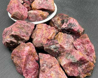 Op de afbeelding: Een verzameling ruwe rhodoniet mineraalspecimens met dieproze en magentakleurige tinten en donkere zwarte mangaankleurige insluitingen. Verschillende grillige, ongepolijste stenen liggen op een hoop, deels in een kleine witte kom.