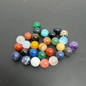 Pode incluir: Uma coleção de pequenas contas de pedras preciosas esféricas e lisas em uma variedade de cores e padrões, incluindo branco, azul, verde, vermelho, amarelo, laranja e roxo sólidos, bem como variedades manchadas, marmorizadas e translúcidas.