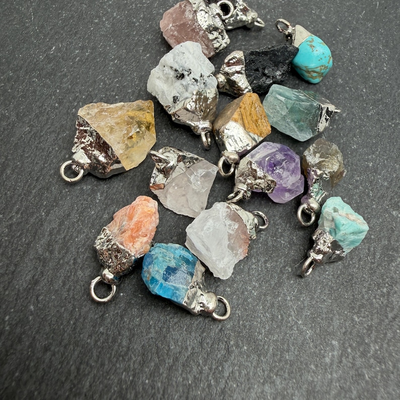 Pu&ograve; includere: Una collezione di ciondoli in pietre preziose grezze e non lucidate in vari colori, tra cui giallo, bianco, nero, blu, viola e arancione. Ogni pietra &egrave; incastonata in una calotta di metallo argentato con un piccolo anello circolare in cima.