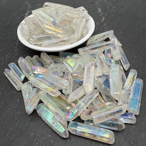 May include: A collection of clear, elongated quartz crystal points with an iridescent, rainbow-like metallic coating. Many of the crystals are piled together on a dark surface, while others are contained within a small, round white ceramic bowl.