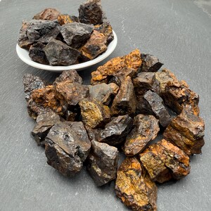 Puede incluir: Una colección de piedras minerales en bruto y dentadas que presentan una mezcla de tonos gris carbón oscuro, negro metálico y naranja parduzco oxidado. Algunas piedras están en un cuenco blanco y otras esparcidas en un montón.