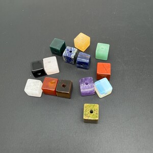 May include: A collection of fifteen small, cube-shaped beads made from various polished stones. Each bead features a single centred hole. The assortment includes colours such as black, white, orange, dark green, blue, red, brown, purple, and light green.