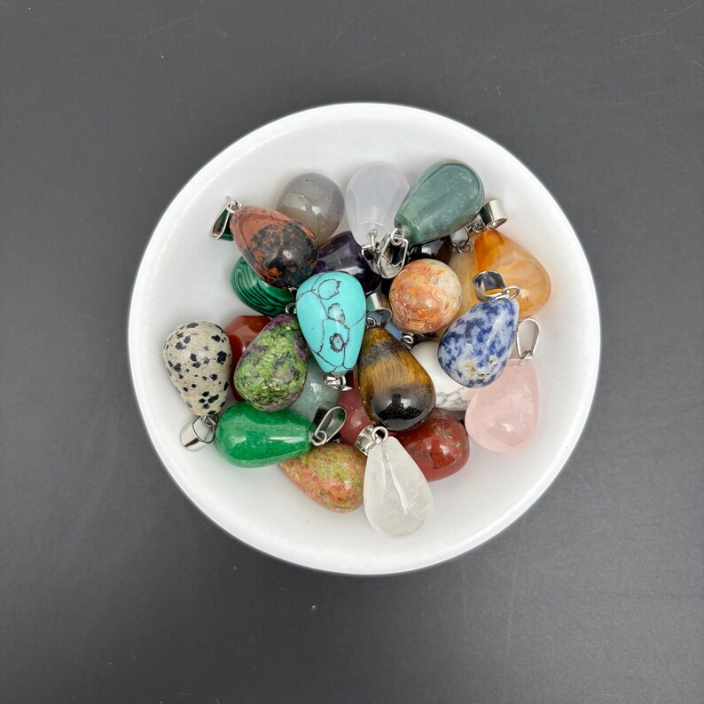 May include: A collection of teardrop-shaped gemstone pendants in a white bowl. The stones feature various colors and patterns, including spotted dalmatian jasper, green malachite, blue turquoise, tiger eye, rose quartz, and clear quartz with metal bails.