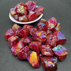May include: A collection of raw, angular crystal chunks with a vibrant magenta and red base color, featuring an iridescent, metallic rainbow coating that reflects gold, blue, and purple hues, with some pieces arranged in a small white bowl.