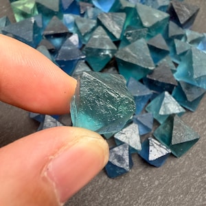 May include: A collection of small, translucent, teal-colored fluorite crystals shaped as octahedrons. Each crystal features a geometric, multi-faceted structure with sharp edges and a smooth, glassy texture, displaying varying shades of blue and green.