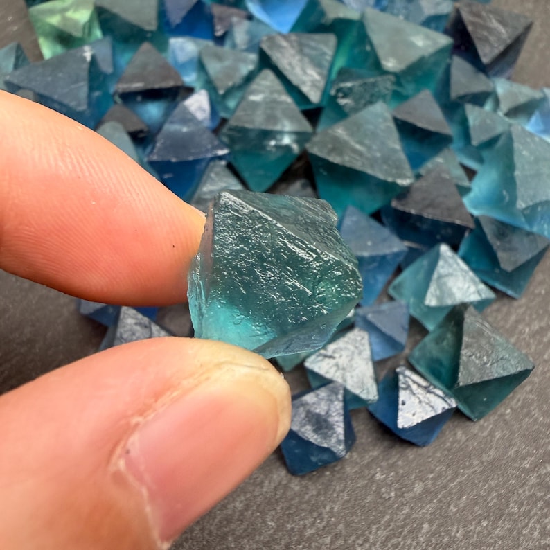May include: A collection of small, translucent, teal-colored fluorite crystals shaped as octahedrons. Each crystal features a geometric, multi-faceted structure with sharp edges and a smooth, glassy texture, displaying varying shades of blue and green.