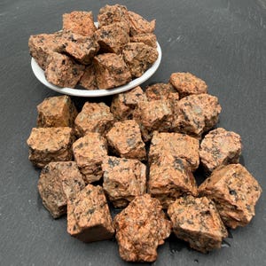 May include: A collection of rough, angular granite stones featuring a mottled texture with shades of salmon pink, tan, and black mineral flecks. Some stones are piled inside a small white bowl, while others are scattered across the surface.