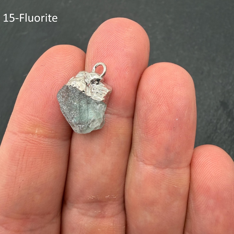 Pu&ograve; includere: Un ciondolo in fluorite verde grezza e traslucida con un cappuccio in metallo color argento e un piccolo anello circolare sulla parte superiore. La pietra ha una forma irregolare e frastagliata, etichettata 15-Fluorite nell'angolo in alto a sinistra.
