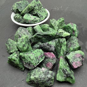 Könnte beinhalten: Eine Sammlung roher Rubin-in-Zoisit-Steine, die eine Mischung aus leuchtend grünen und tief magentafarbenen Mineralablagerungen mit schwarzen Sprenkeln aufweisen. Einige Steine befinden sich in einer kleinen weißen Schale, andere liegen verstreut.
