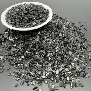 May include: A white ceramic bowl filled with small, polished black stone chips, with additional black stone chips scattered around the base of the bowl. The stones have a glossy, reflective surface and irregular, angular shapes throughout.