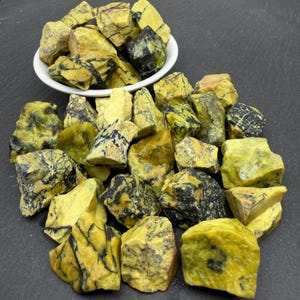 Puede incluir: Una colección de piedras en bruto y sin pulir con una base de color amarillo vibrante y vetas negras oscuras muy marcadas. Algunas de estas piezas minerales irregulares y dentadas están en un cuenco blanco, mientras otras están dispersas.