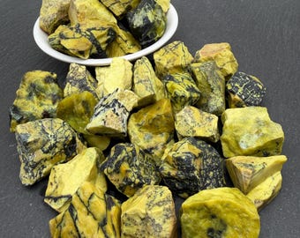 Op de afbeelding: Een verzameling ruwe, ongepolijste stenen met een levendige gele basiskleur en opvallende, donkerzwarte aders. Sommige van deze grillige, onregelmatige mineraalstukken zitten in een witte kom, terwijl andere los verspreid liggen.