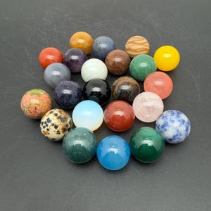 May include: A collection of twenty-one smooth, spherical stones in various colours and textures, including solid yellow, red, blue, green, black, white, and translucent pink, alongside speckled, marbled, and glittery mineral varieties.