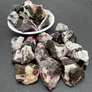 May include: A collection of raw, unpolished mineral stones featuring a mix of dark charcoal grey, stark white, and vibrant crimson red coloration. Some stones are contained within a small white bowl, while others are scattered around the base.