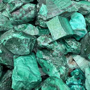Op de afbeelding: Een verzameling ruwe malachietstenen met diepe bosgroene en levendige smaragdgroene tinten. De mineraalspecimens vertonen natuurlijke banden, gelaagde texturen en grillige, kristallijne oppervlakken in verschillende groentinten.