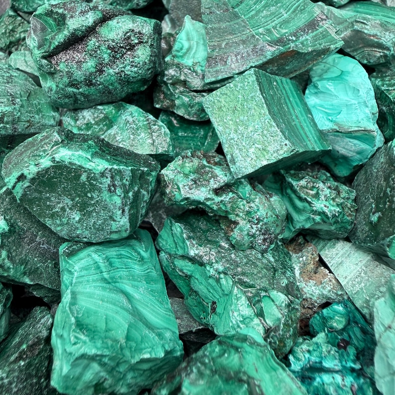 Op de afbeelding: Een verzameling ruwe malachietstenen met diepe bosgroene en levendige smaragdgroene tinten. De mineraalspecimens vertonen natuurlijke banden, gelaagde texturen en grillige, kristallijne oppervlakken in verschillende groentinten.
